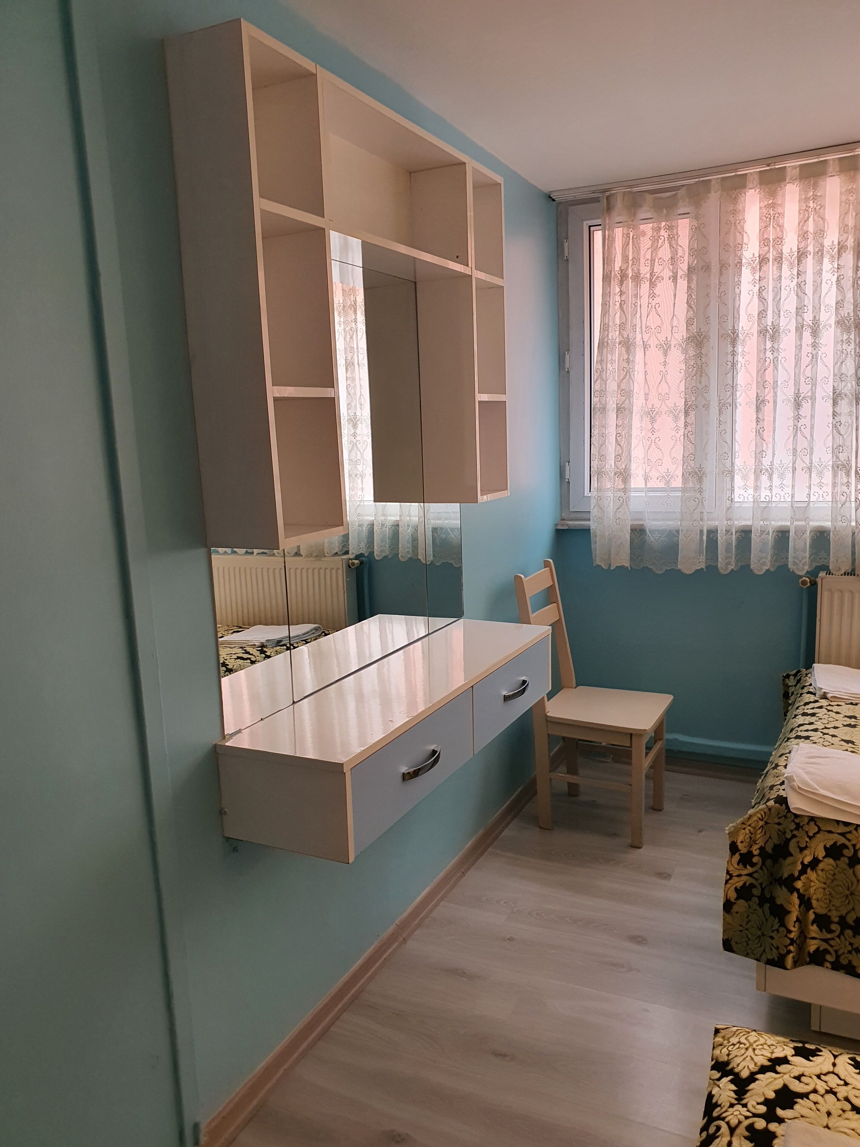 A bedroom with a large mirror, a wooden chair, a bed with patterned bedding, and a window with sheer curtains.