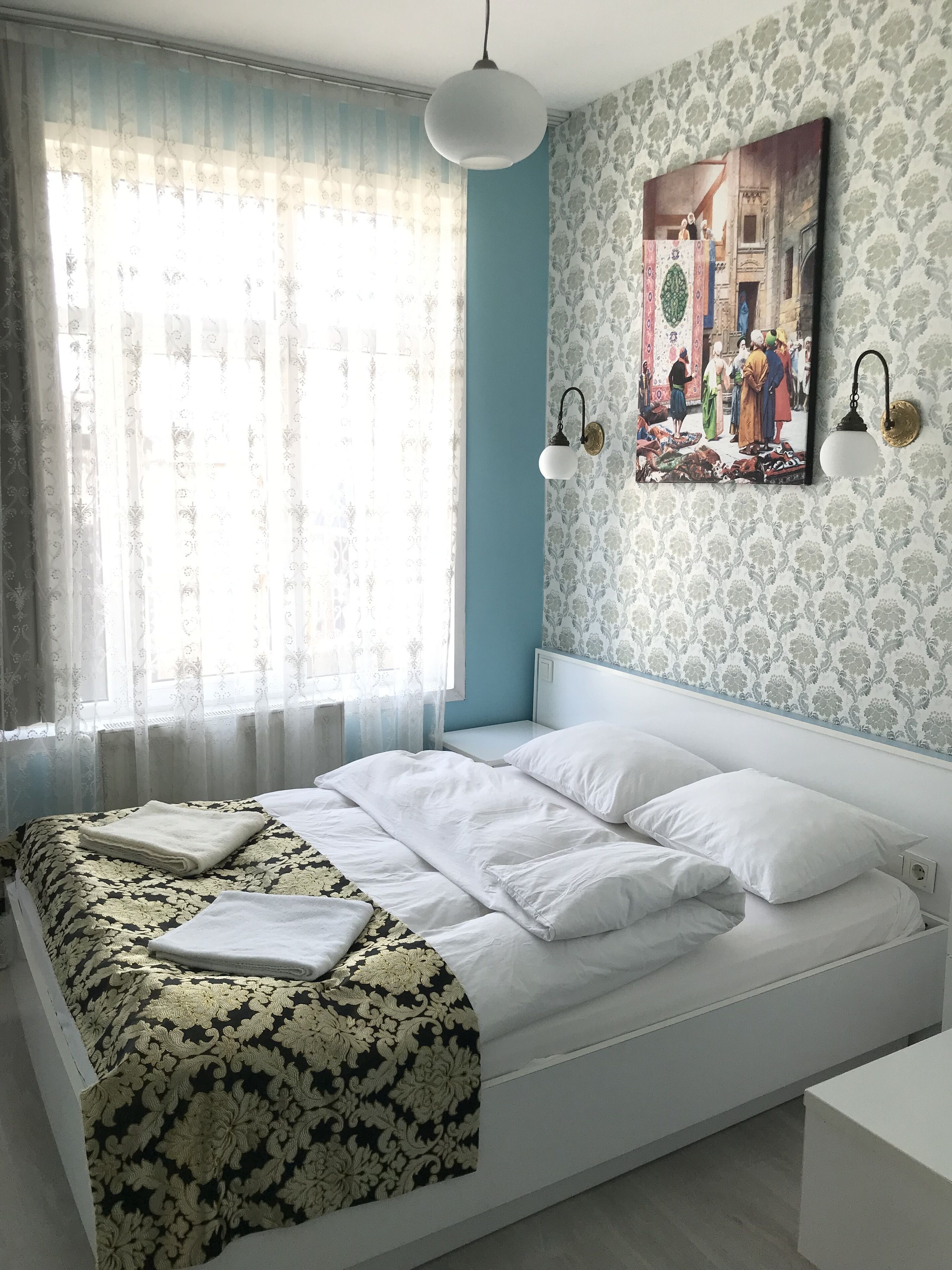A bedroom with a large bed, a bedside table, a wall-mounted lamp, and a window with sheer curtains.
