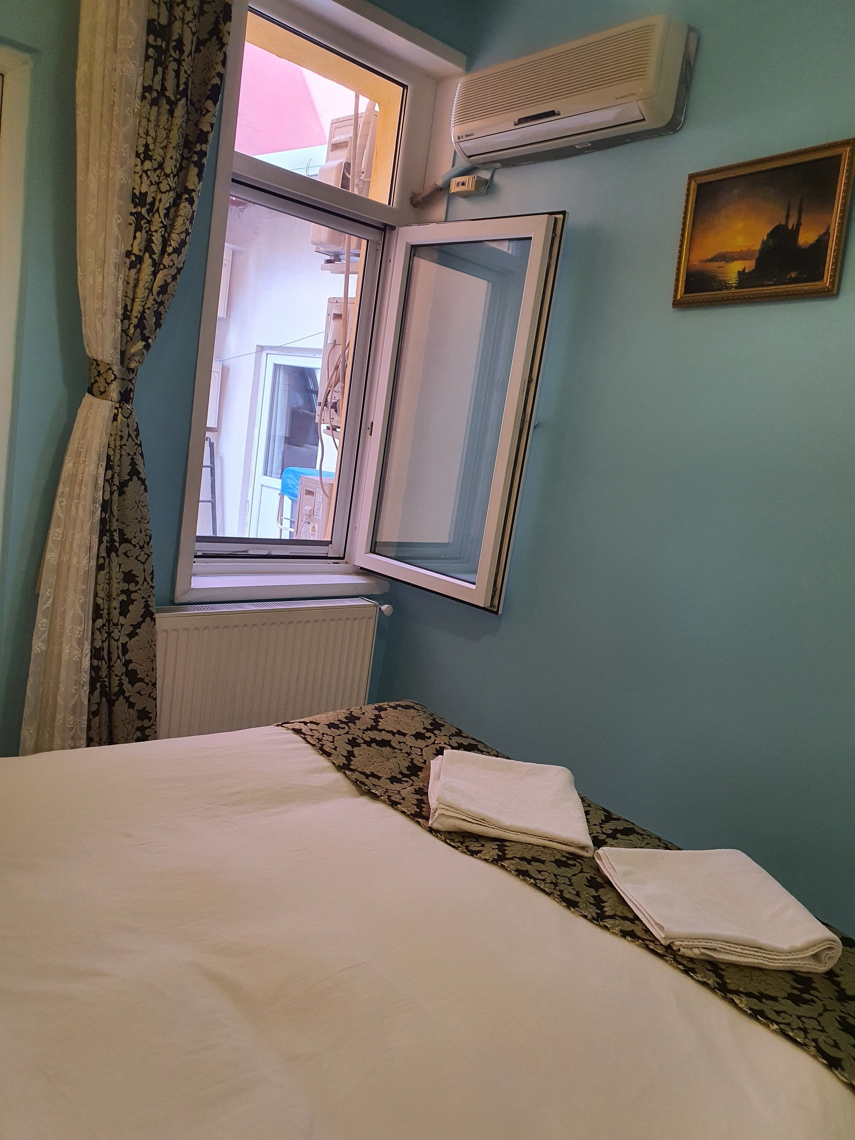 A bedroom with a bed, a window, an air conditioner, and a painting on the wall.