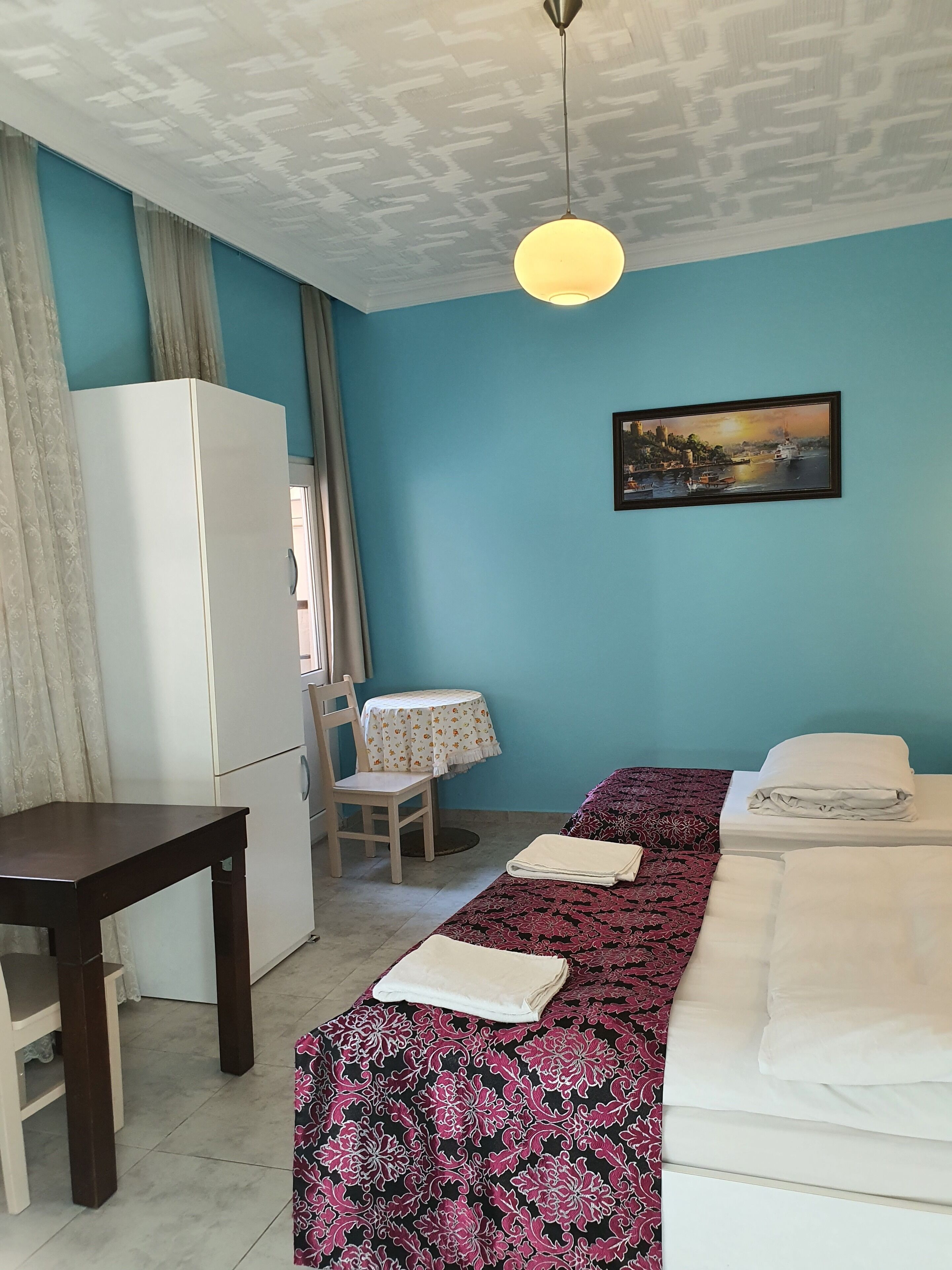 Una habitación con cama, escritorio, silla, mesa, refrigerador y un cuadro en la pared.