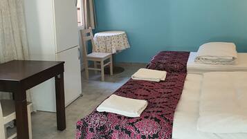 Una habitación con cama, escritorio, silla, mesa, refrigerador y un cuadro en la pared.