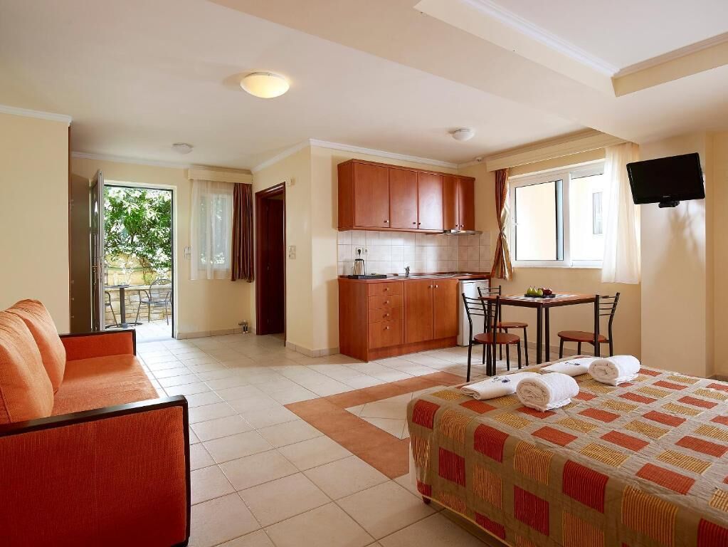 A hotel room with a bed, a sofa, a dining table, a kitchen area, and a TV.