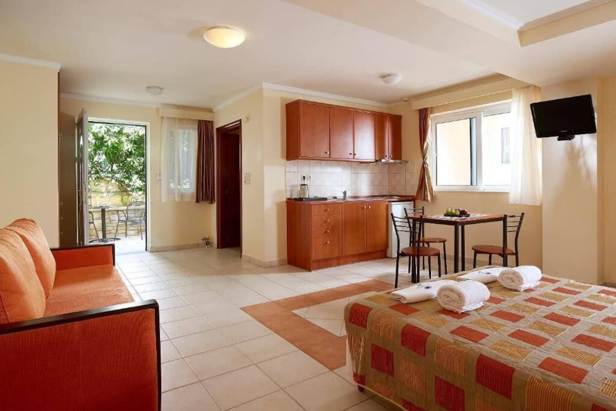 A hotel room with a bed, a sofa, a dining table, a kitchen area, and a TV.