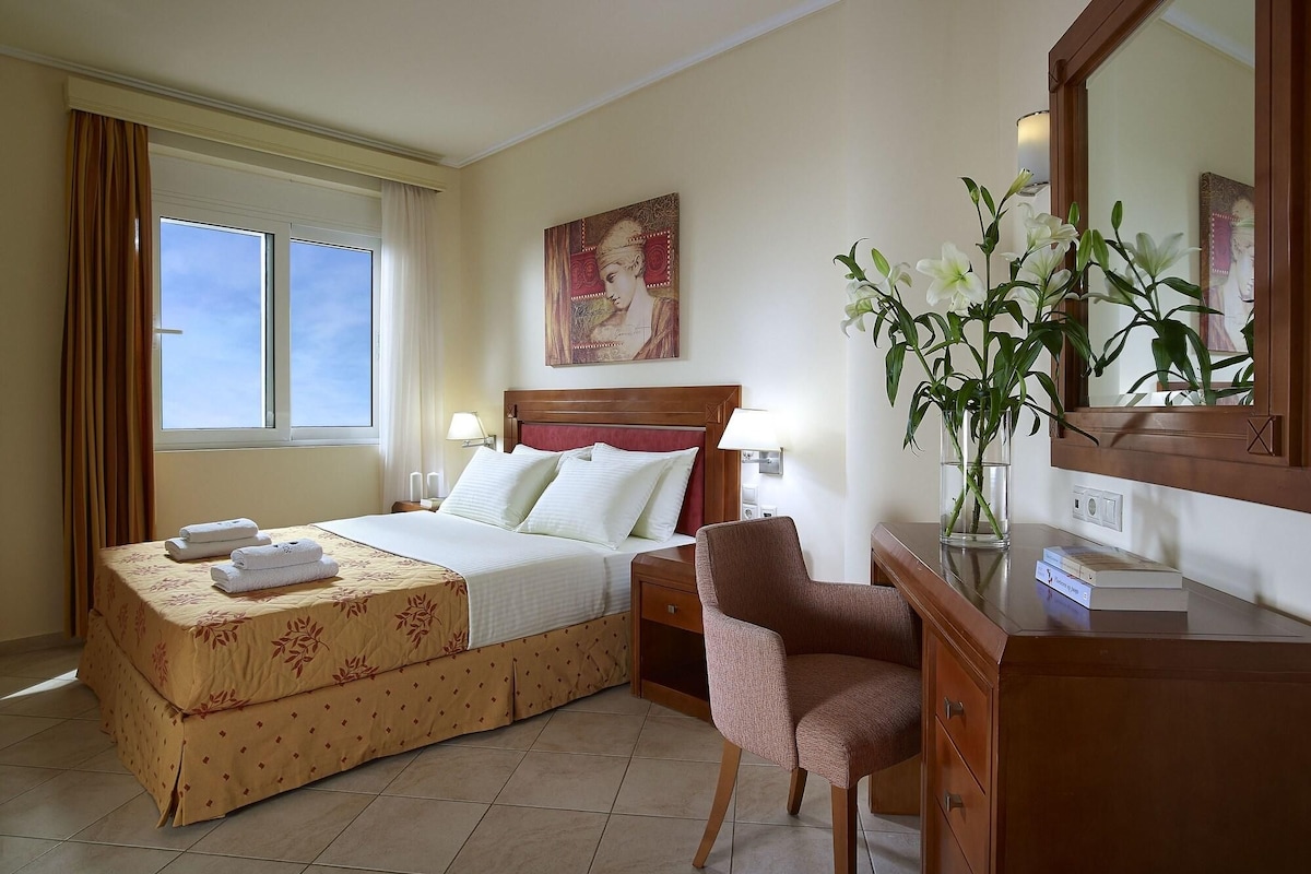 A hotel room with a bed, a chair, a desk, a lamp, a vase with flowers, a painting on the wall, and a window with curtains.