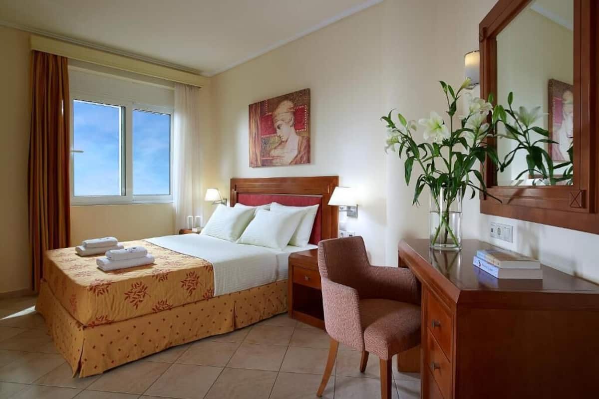 A hotel room with a bed, bedside tables, a chair, a desk, a mirror, and a window.
