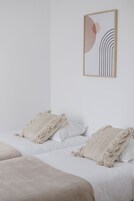 A minimalist bedroom with two beds, each with a white mattress and a beige blanket. A framed abstract artwork hangs on the wall above the beds.