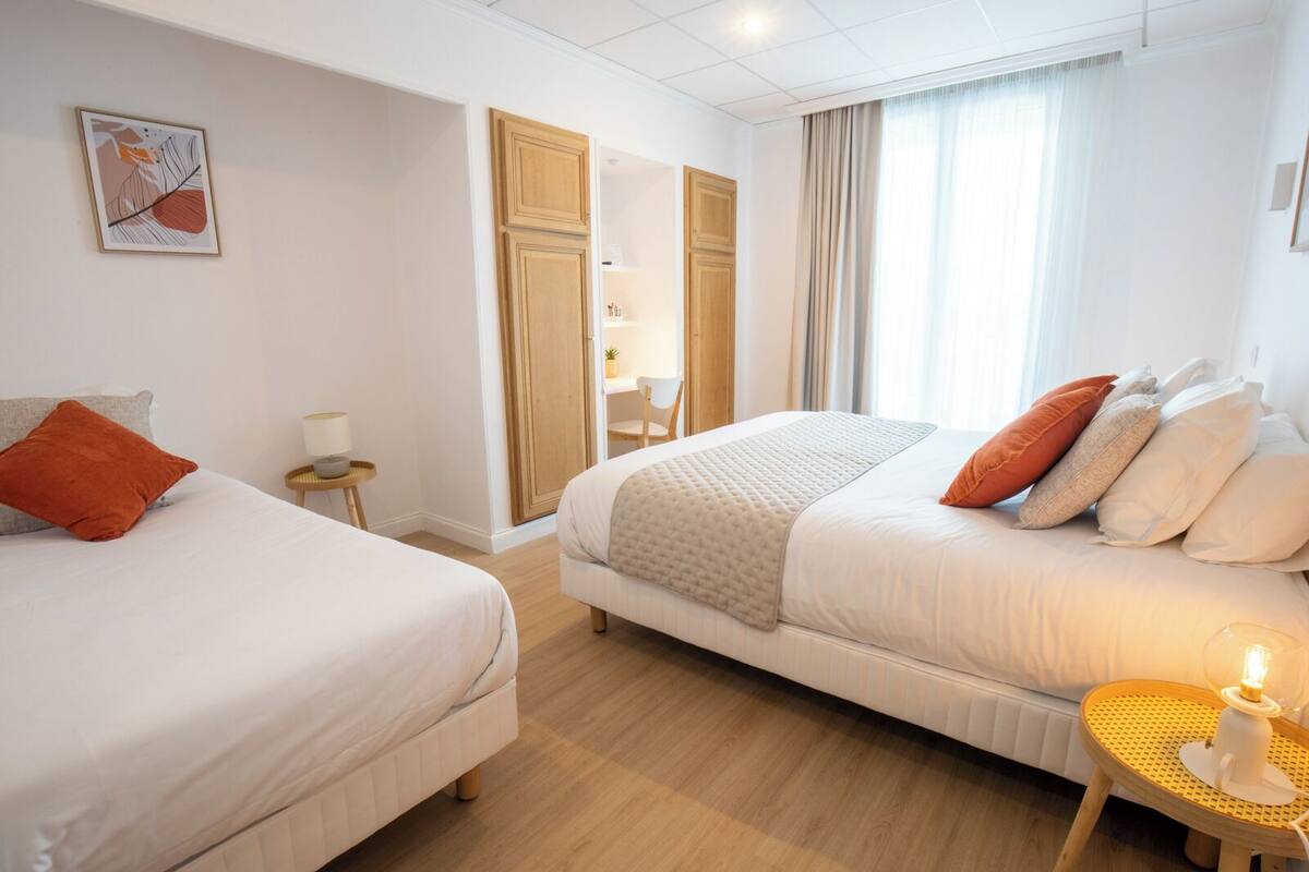 A hotel room with two beds, a small table with a lamp, a painting on the wall, and a window with curtains.