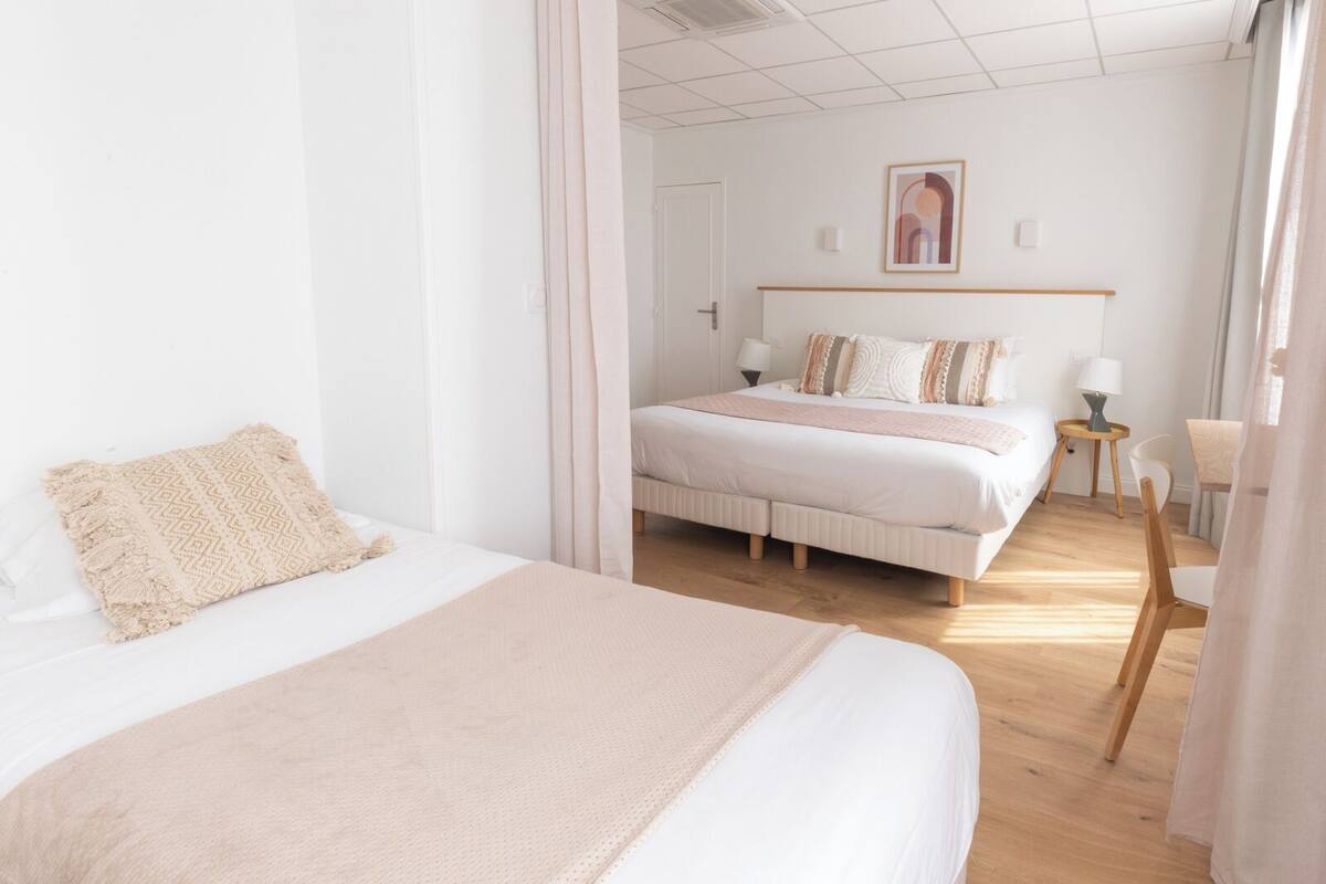 A hotel room with two beds, a wooden floor, a painting on the wall, and a small table with a lamp.