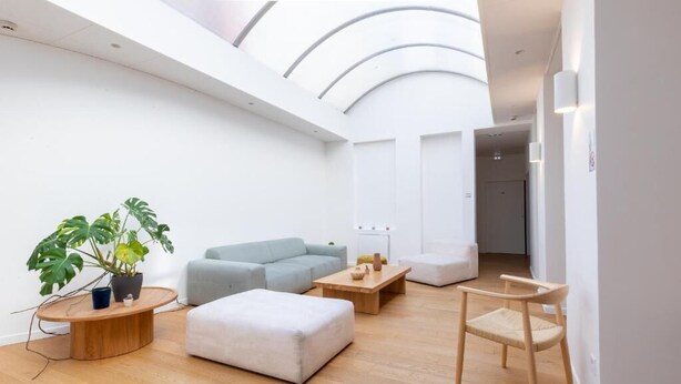 A modern living room with a high ceiling, a light blue sofa, a white ottoman, a wooden coffee table, and a wooden chair. - Hôtel Porte Mars Reims Gare Centre (Reims)