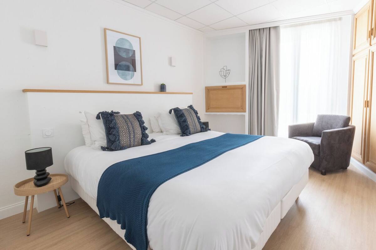 A hotel room with a large bed, a bedside table with a lamp, a chair, and a window with curtains.