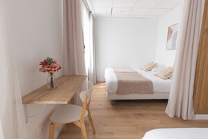A bedroom with a wooden desk, a chair, a bed with a beige blanket, and a vase with flowers.