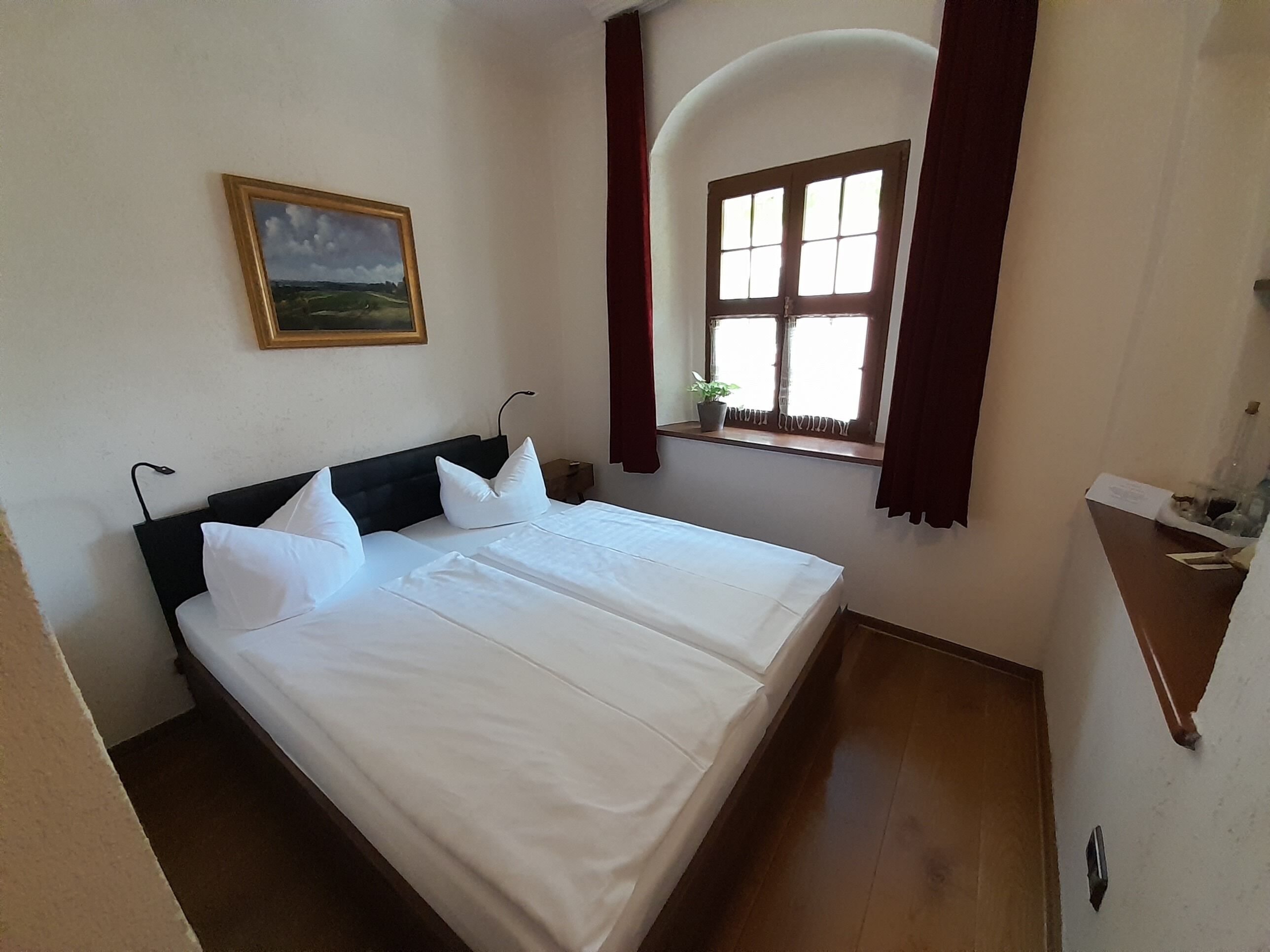 Un lit bien fait, avec du linge de lit blanc et des oreillers, une petite table de chevet avec une lampe, une fenêtre avec des rideaux et un tableau accroché au mur.