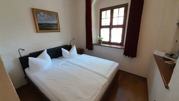 Un lit bien fait, avec du linge de lit blanc et des oreillers, une petite table de chevet avec une lampe, une fenêtre avec des rideaux et un tableau accroché au mur.