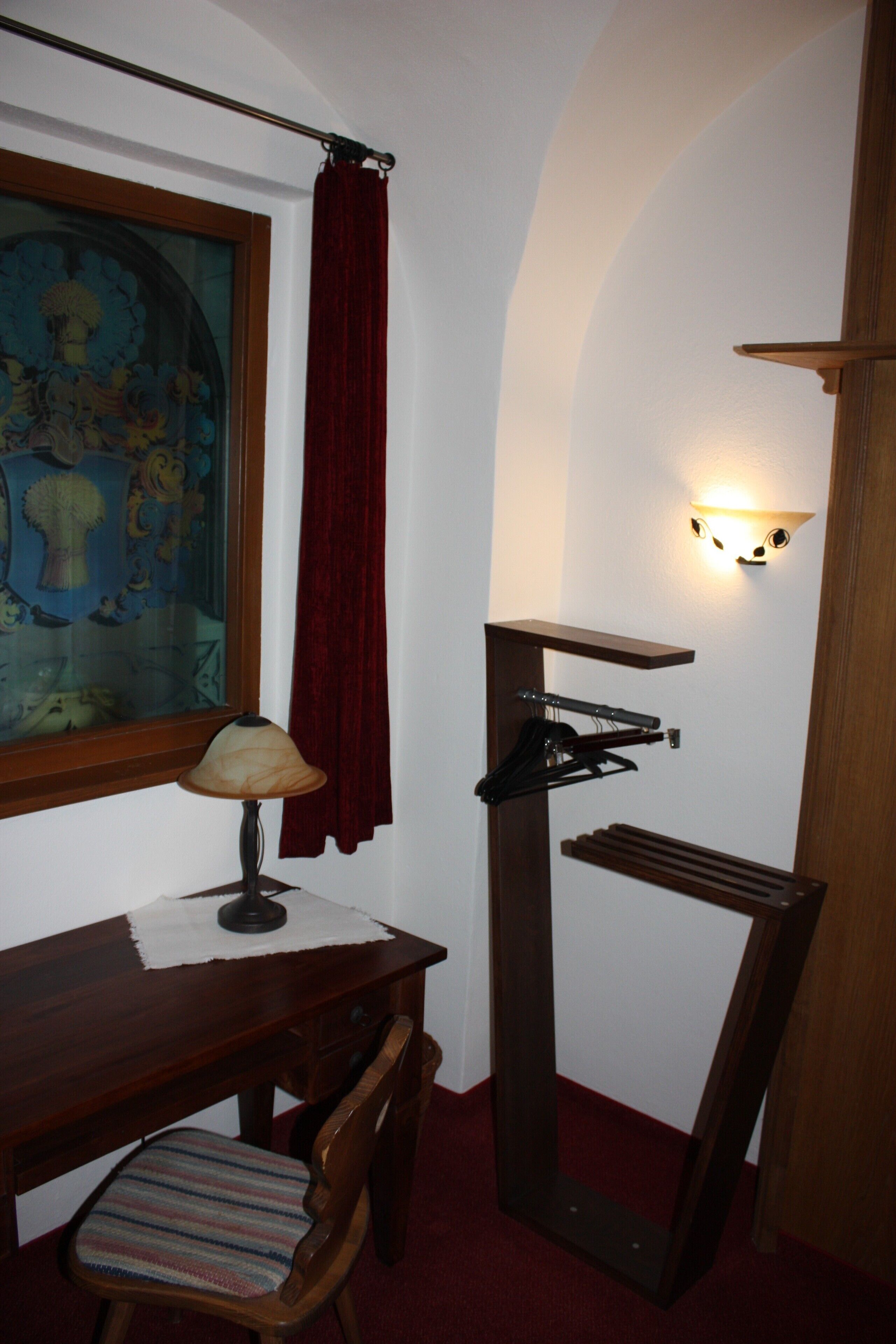 Una habitación con un escritorio de madera, una silla, una lámpara, un perchero y un cuadro en la pared.