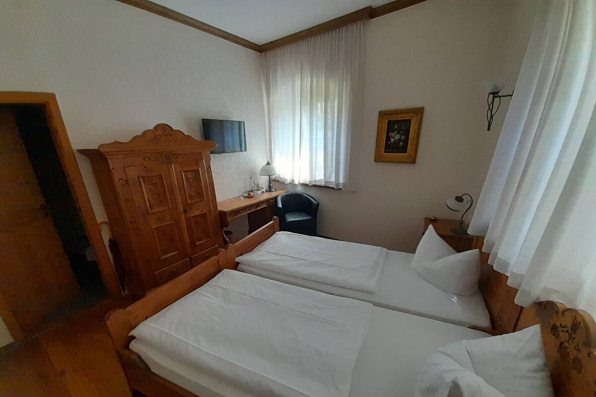 A room with two wooden beds, a desk, a chair, a wardrobe, a window with curtains, and a wall-mounted picture.