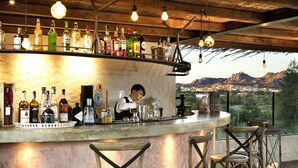 Un bar con una variedad de botellas y vasos, sillas de madera y una vista de montañas al fondo.