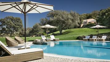Zona de piscina con sillones de descanso y sombrillas, rodeada por un jardín ajardinado y una ladera.