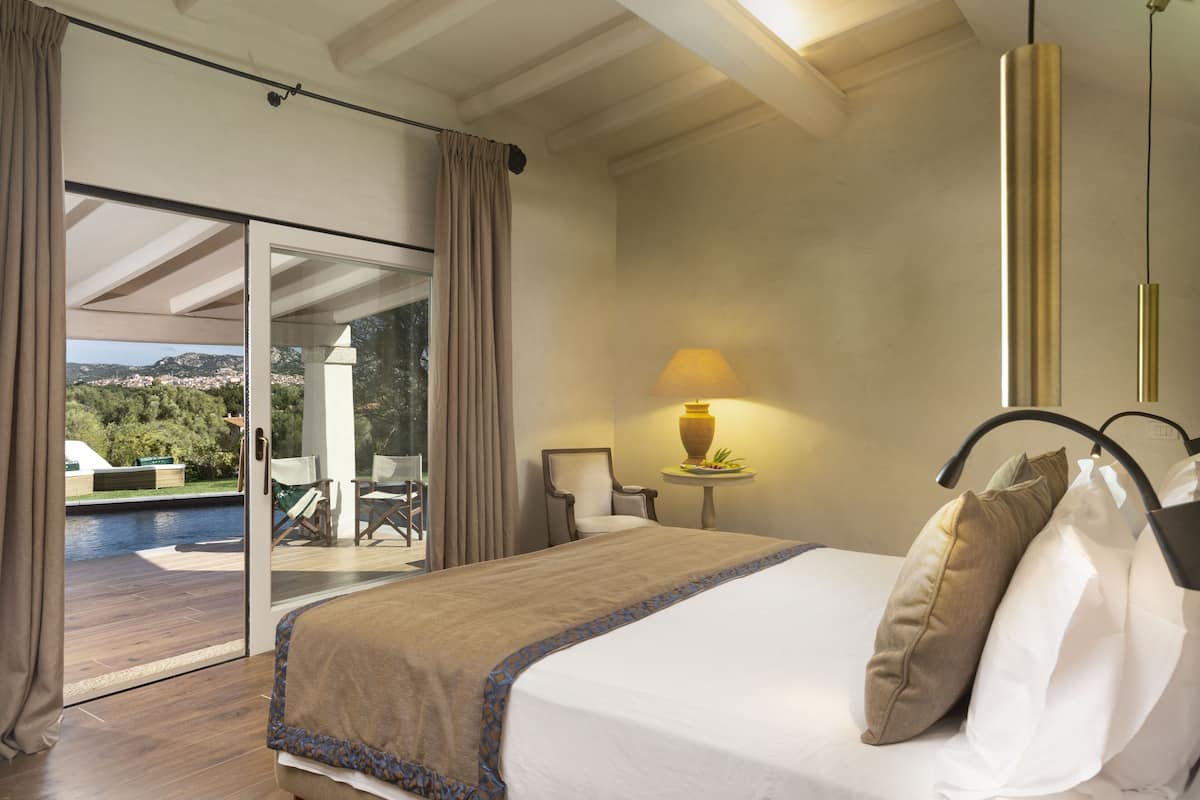 A bedroom with a large bed, a view of a pool and outdoor seating, and a lamp on a small table.