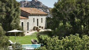 Un edificio de piedra con tejado de tejas, piscina y abundante vegetación.
