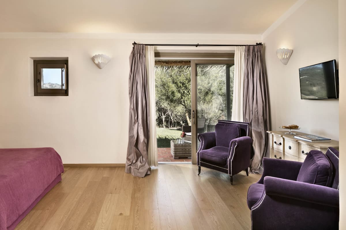 A hotel room with a bed, two armchairs, a TV, a small table with a book and a glass, and a window with curtains.