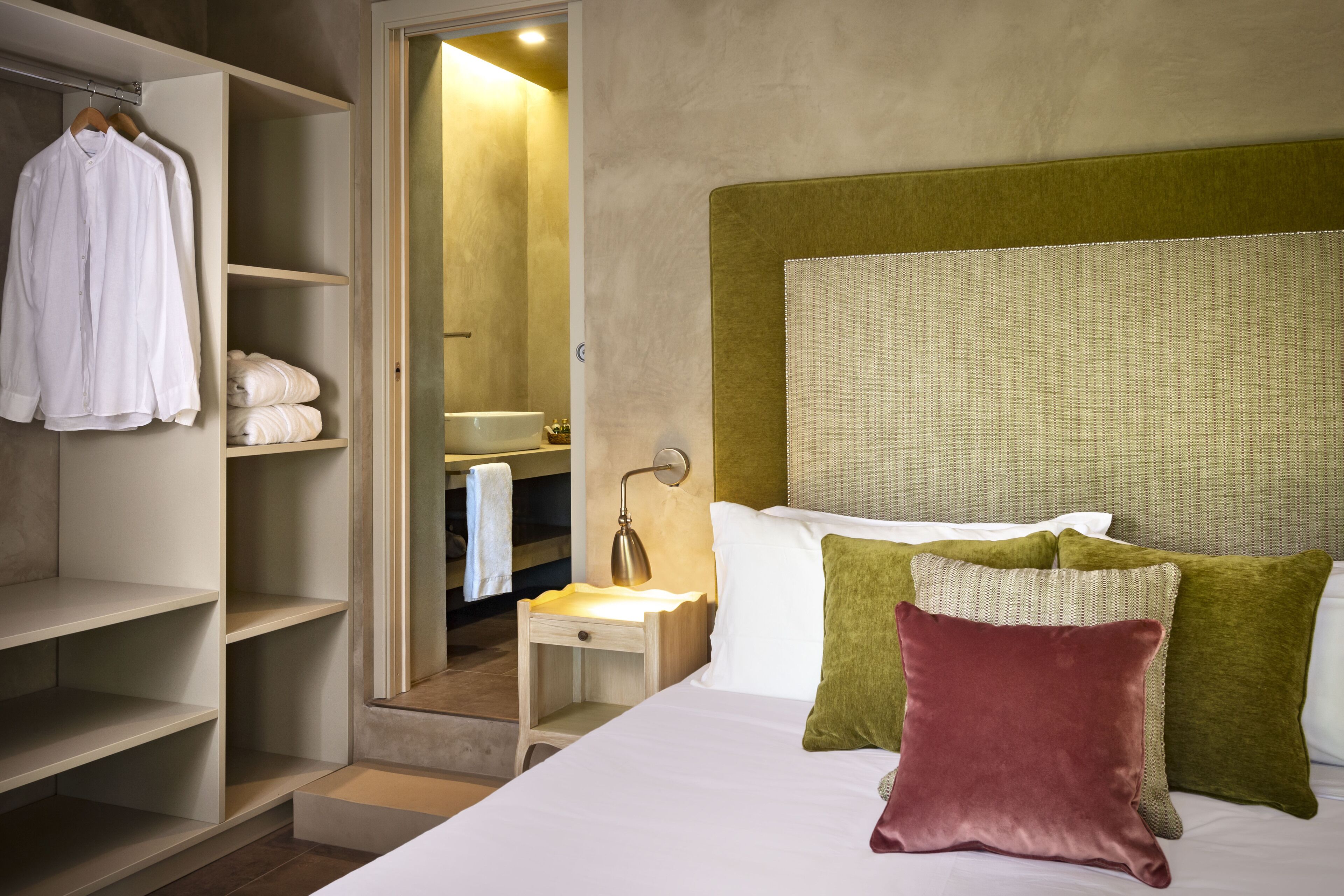 A hotel room with a bed, a nightstand, a bedside lamp, a wardrobe, and a bathroom visible through an open door.