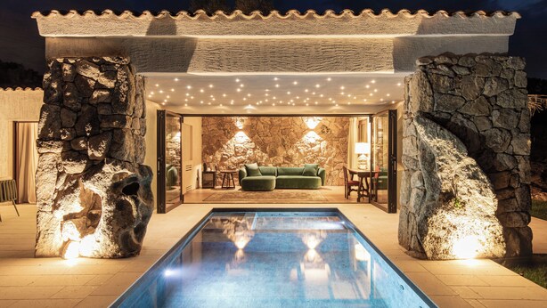 Zona de piscina con muro de piedra, sofá verde e iluminación con luces colgantes.