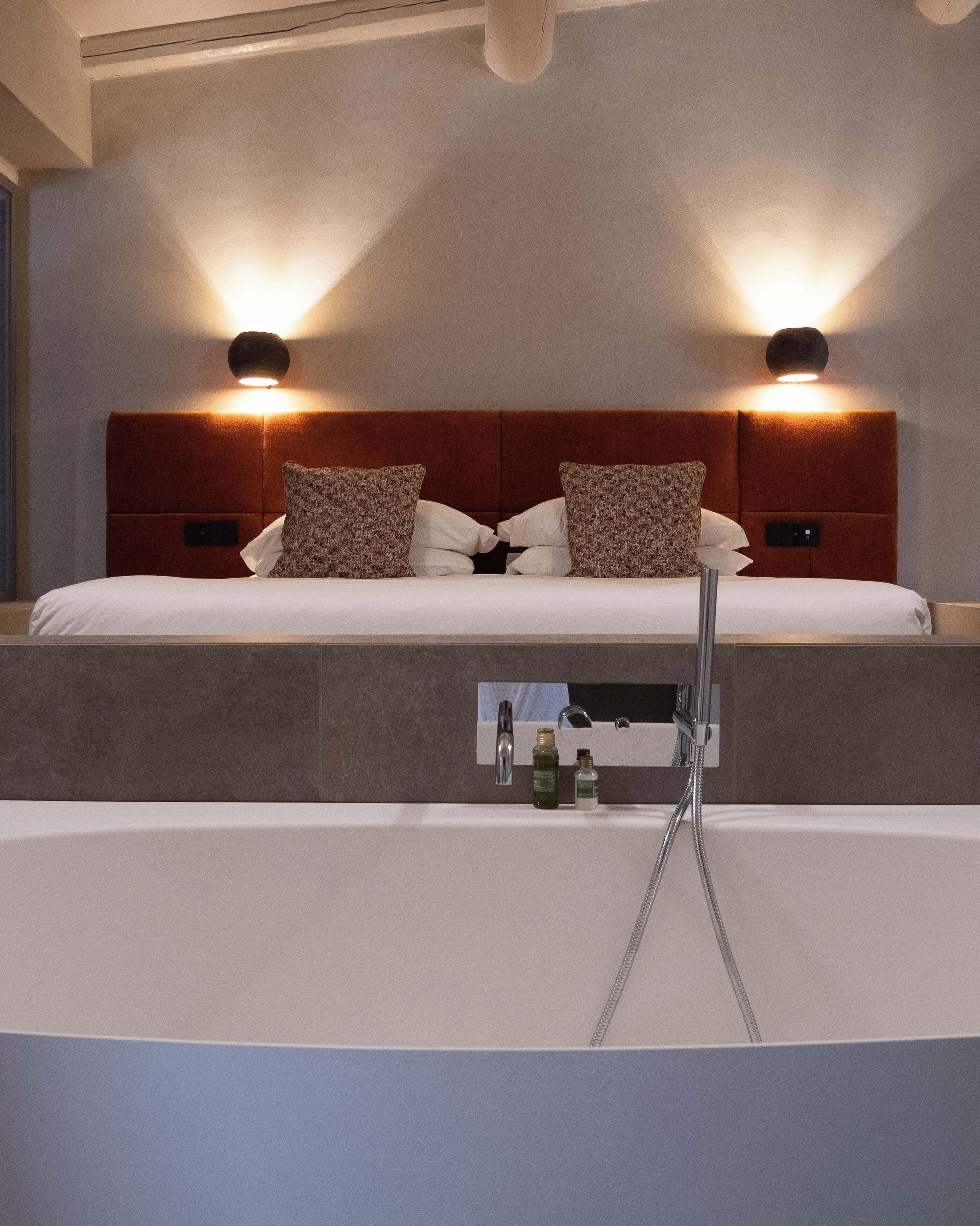A bedroom with a bed, two wall-mounted lights, a headboard, and a built-in bathtub with a showerhead.