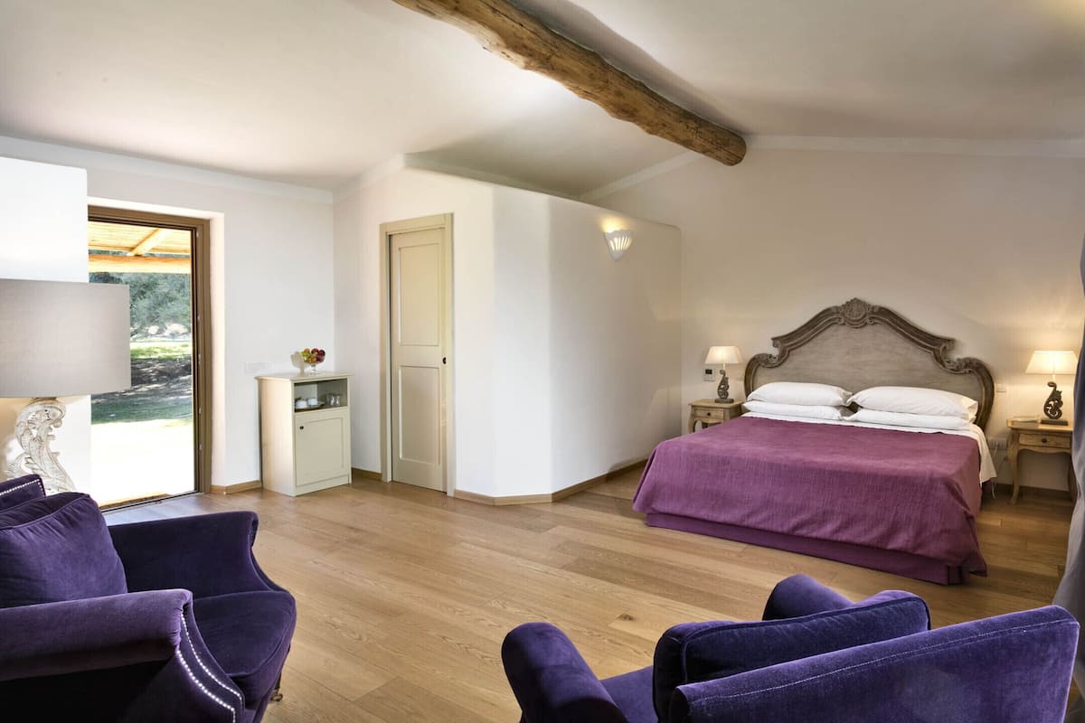 A bedroom with a wooden ceiling, a bed with a purple bedspread, two bedside tables with lamps, a nightstand, a chair, and a small cabinet.