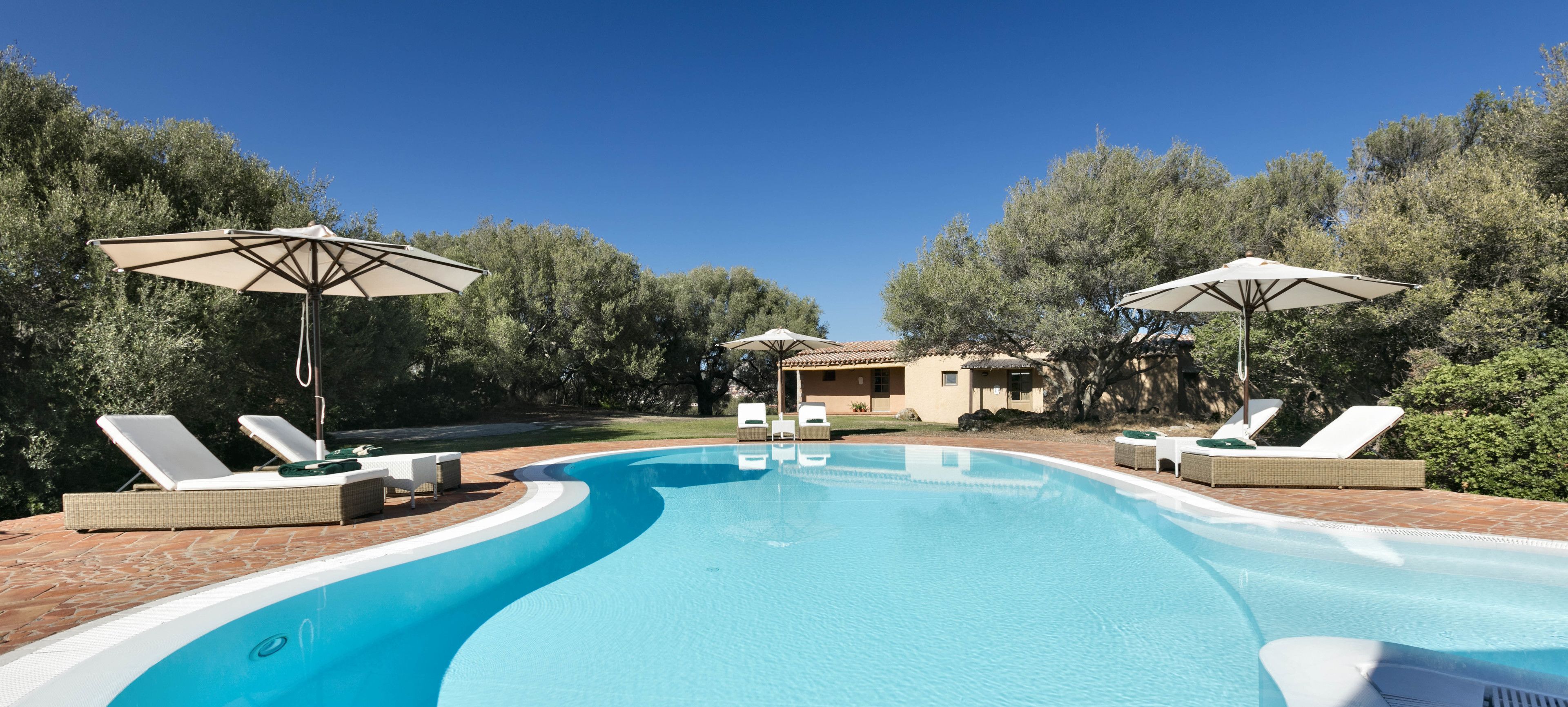 Una piscina con sillones de descanso y sombrillas, rodeada de árboles y una casa al fondo.