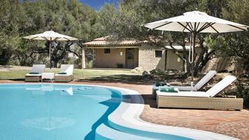 Piscina com espreguiçadeiras e guarda-sóis, cercada por um pátio de pedra e um edifício com telhado telhado.