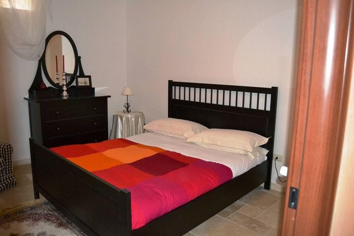 A bedroom with a black bed frame, a colorful quilt, a small bedside table with a lamp, and a mirror on a dresser.