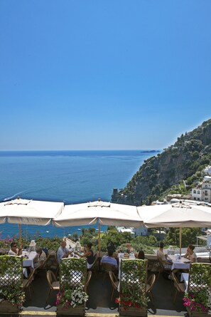 An outdoor dining area with tables and chairs, overlooking a coastal view with a clear sky.