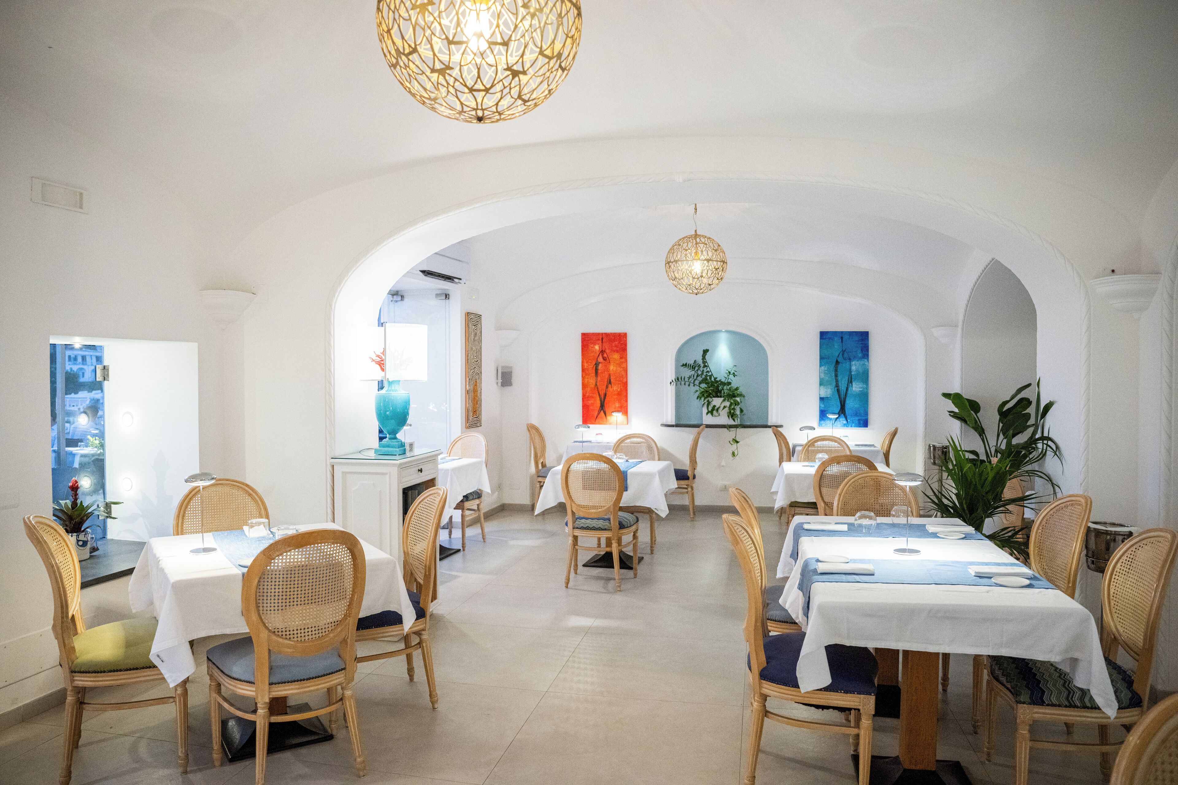 Un restaurante con paredes blancas, muebles de madera y un arco central. Las mesas están cubiertas con manteles blancos y servilletas azules.