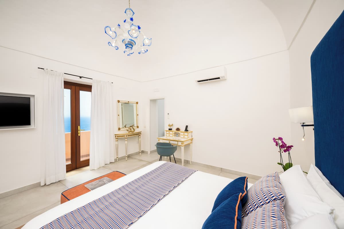 A modern hotel room with a large bed, a desk, a chair, a TV, and a view of the sea through a door.