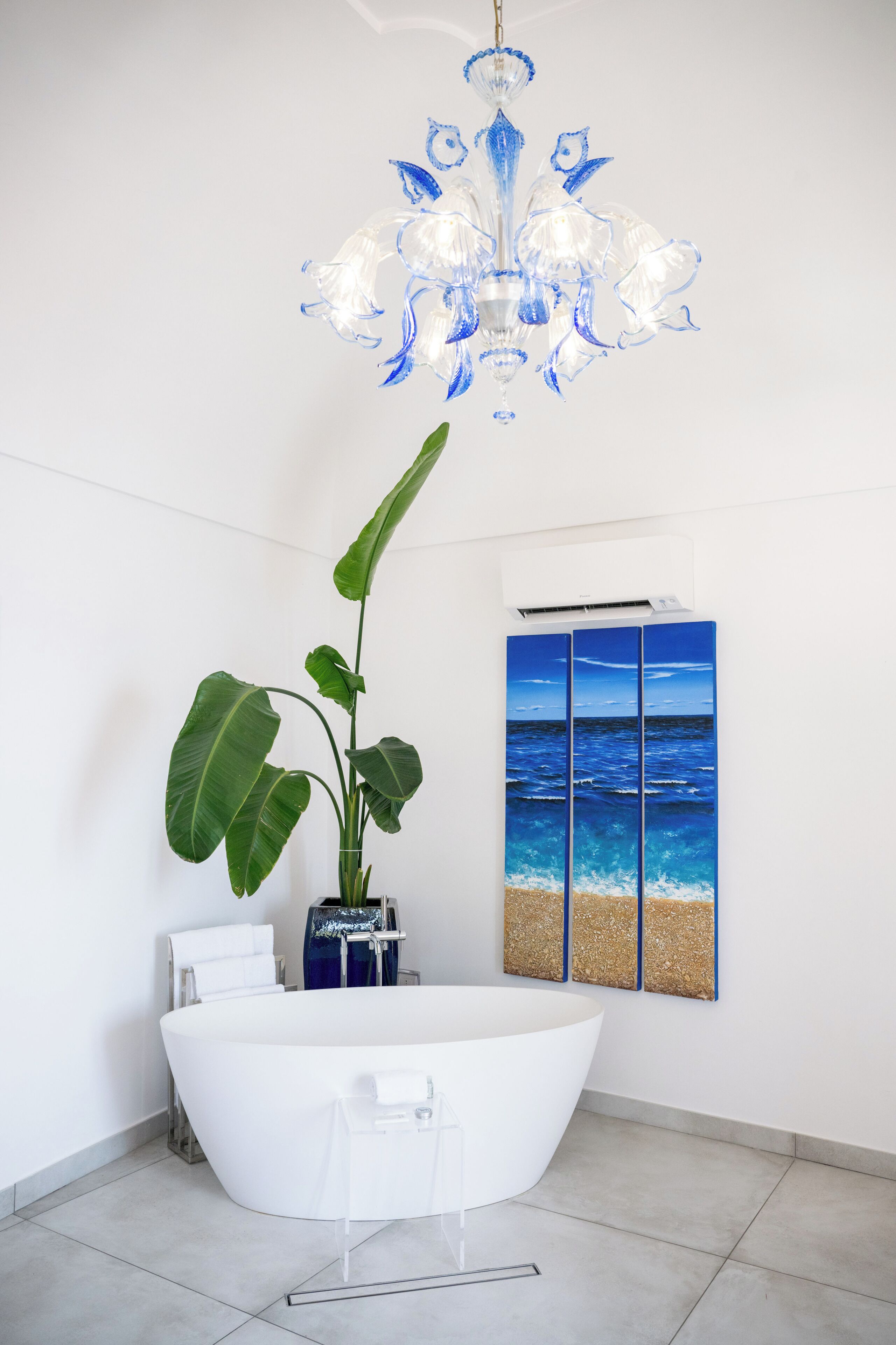A modern bathroom with a freestanding bathtub, a large potted plant, a wall-mounted air conditioner, and a painting of the ocean.