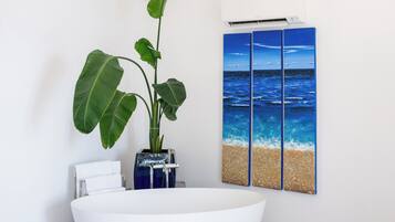 A modern bathroom with a freestanding bathtub, a large potted plant, a wall-mounted air conditioner, and a painting of the ocean.