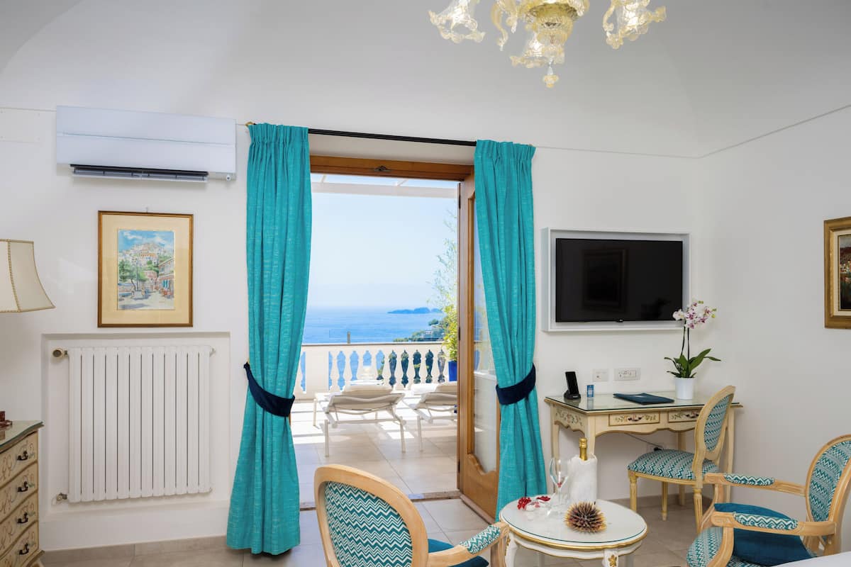 A room with a view of the sea, a desk with a computer, and a dining area with chairs.