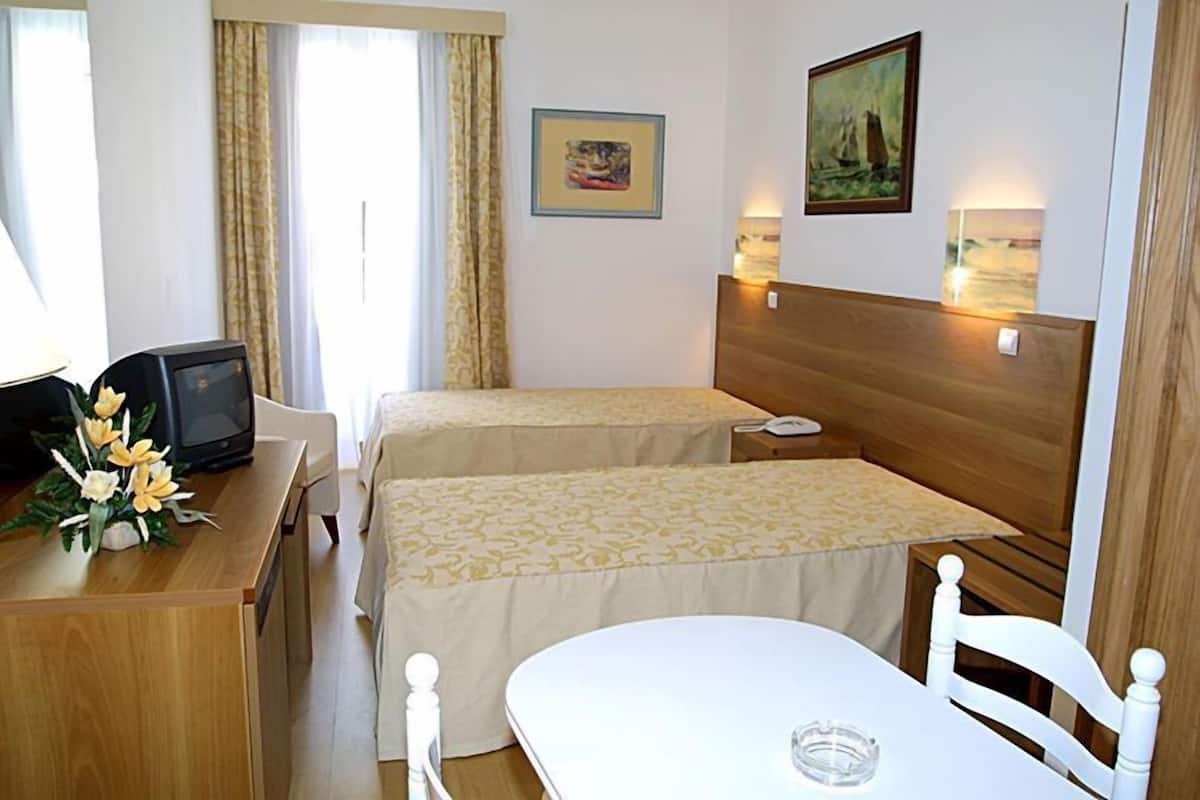 A hotel room with two single beds, a wooden desk with a computer, a small table, and a chair. There are wall-mounted lamps and a painting above the bed.