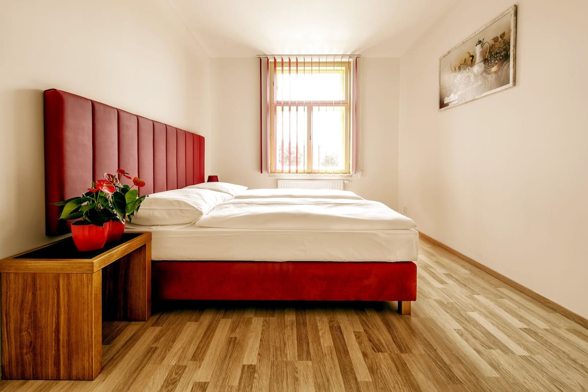 A bedroom with a wooden floor, a red headboard, a white bed, a nightstand with a red vase, and a window with blinds.