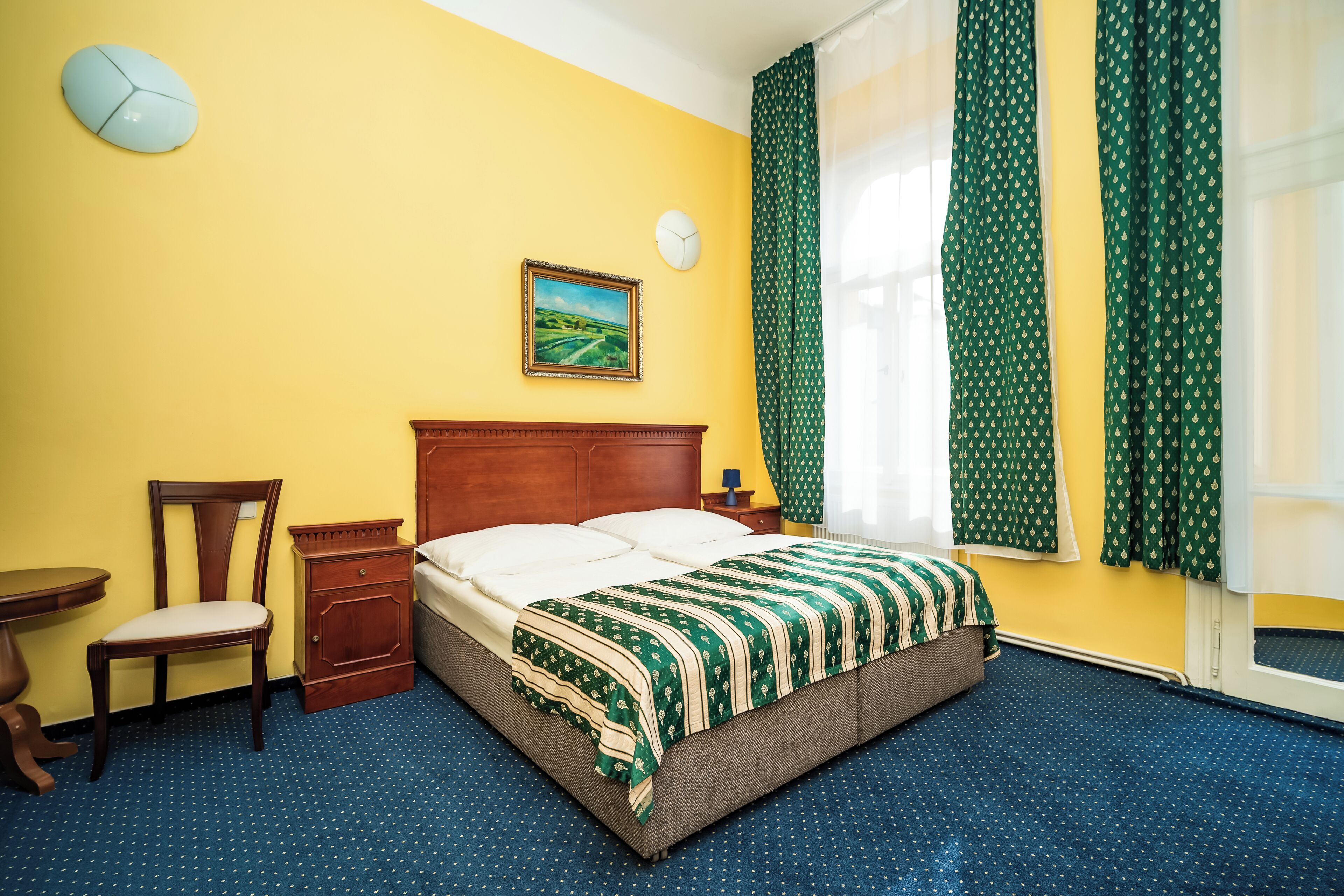 A hotel room with a bed, a chair, a nightstand, a clock, and a painting on the wall.