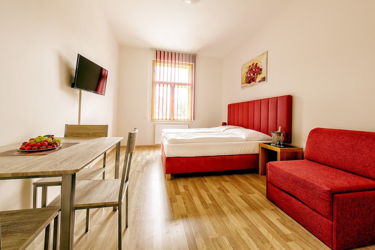 A hotel room with a wooden floor, a bed with a red headboard, a red sofa, a dining table with chairs, and a television on the wall.