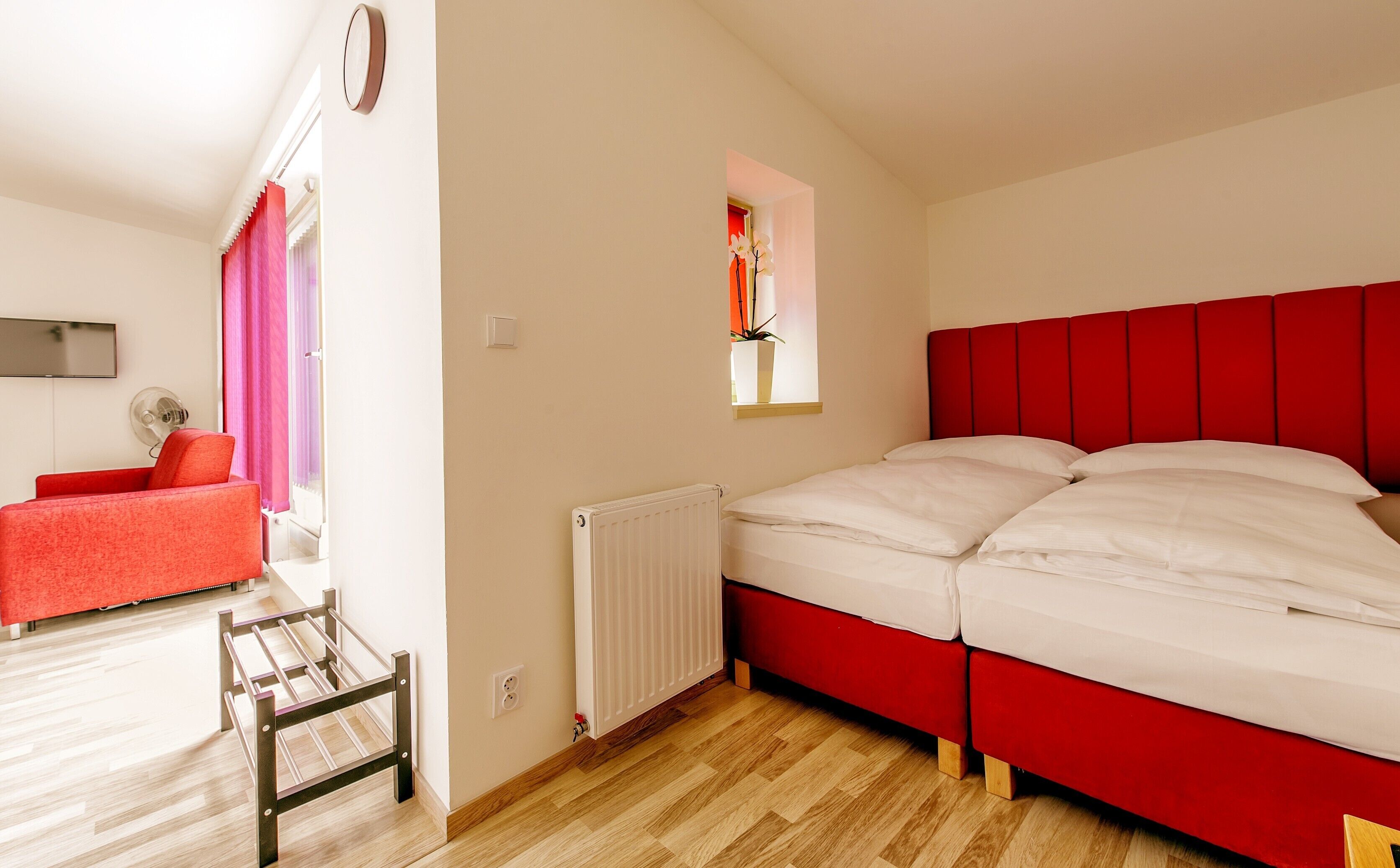 A bedroom with a red headboard, a white bed, a wooden floor, a red chair, a small window with a plant, and a radiator.