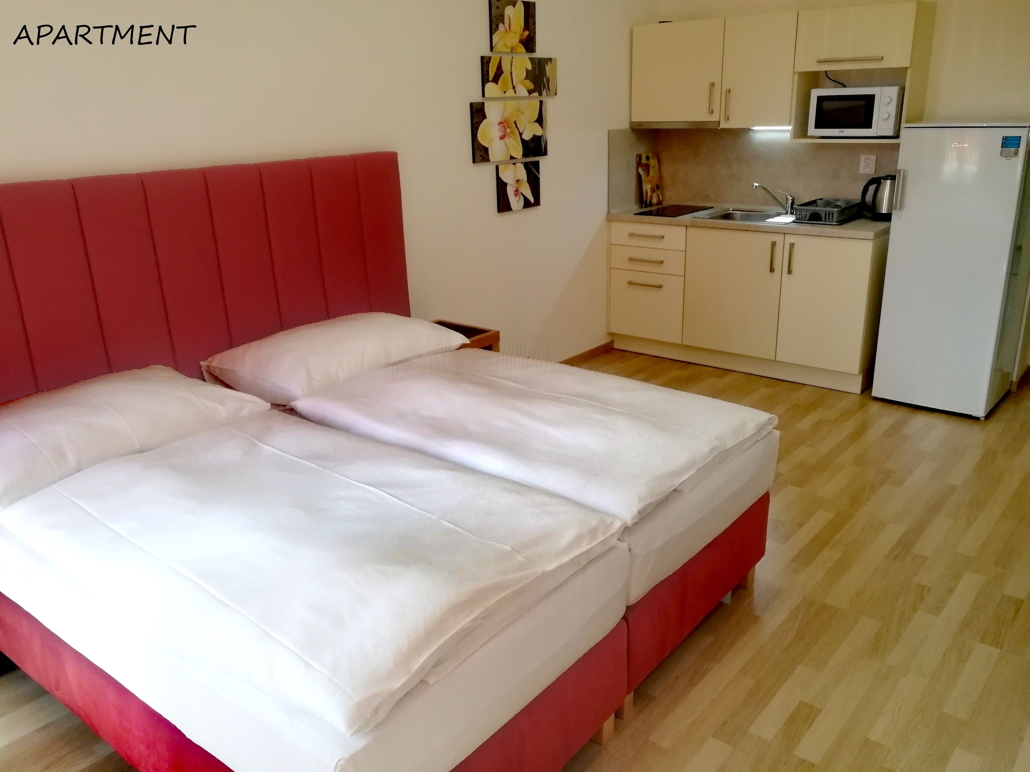 Un dormitorio con cabecera roja, una cama con ropa blanca, una mesita de noche pequeña y una mesilla auxiliar. Junto al dormitorio, hay una kitchenette con microondas, hervidor y refrigerador.