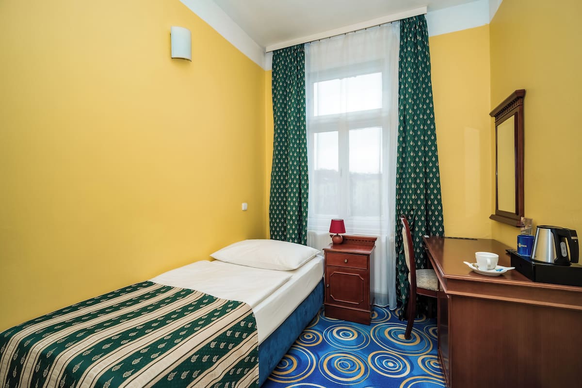A hotel room with a single bed, a wooden nightstand with a lamp, a mirror, and a small table with a cup and a kettle.