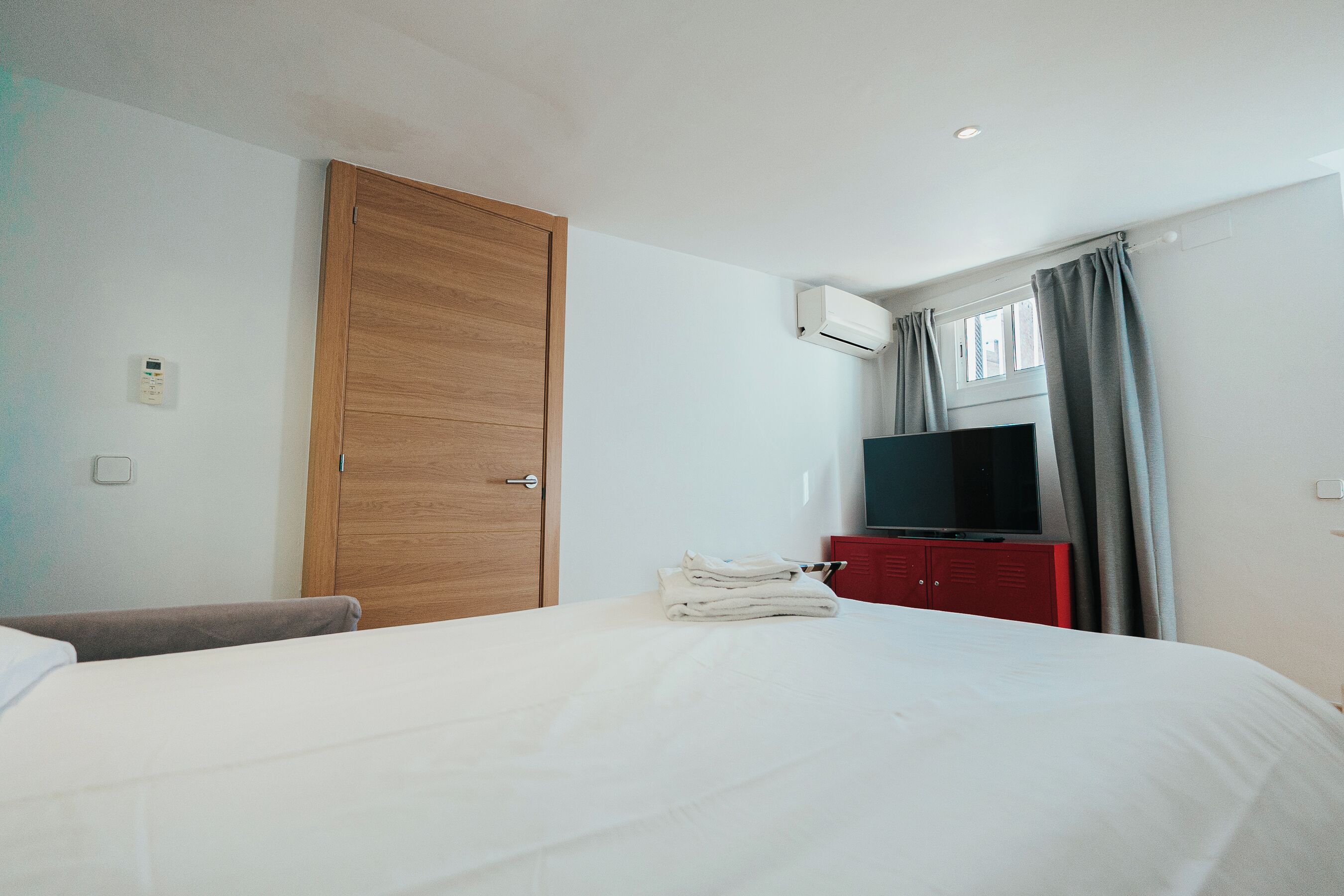 A hotel room with a bed, a wooden wardrobe, a red dresser, a television, and a window with curtains.