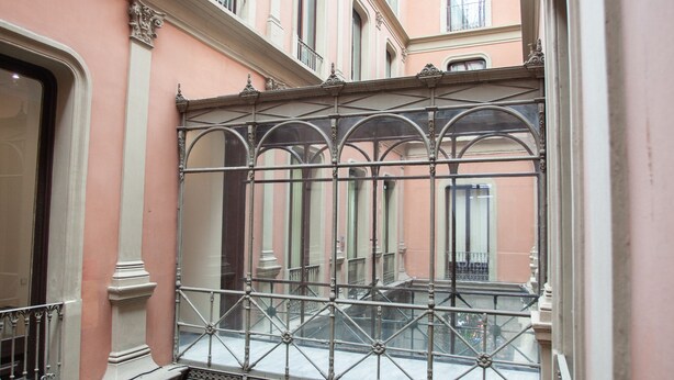 Een smalle gang met decoratieve ijzeren hekwerk en een glazen balustrade, die uitkomt op een balkon met uitzicht op de lucht.