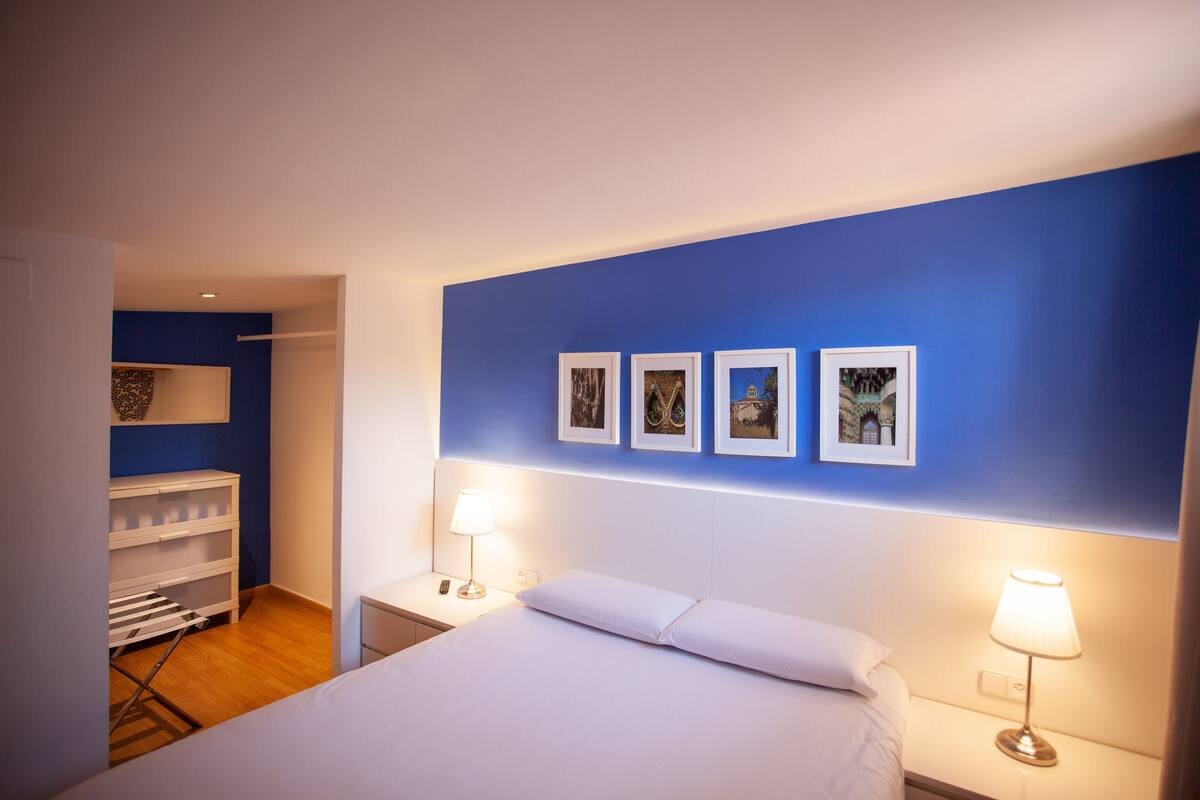 Une chambre d’hôtel avec un lit, deux lampes de chevet, une table de chevet, une commode blanche, un étendoir et quatre tableaux encadrés accrochés au mur.