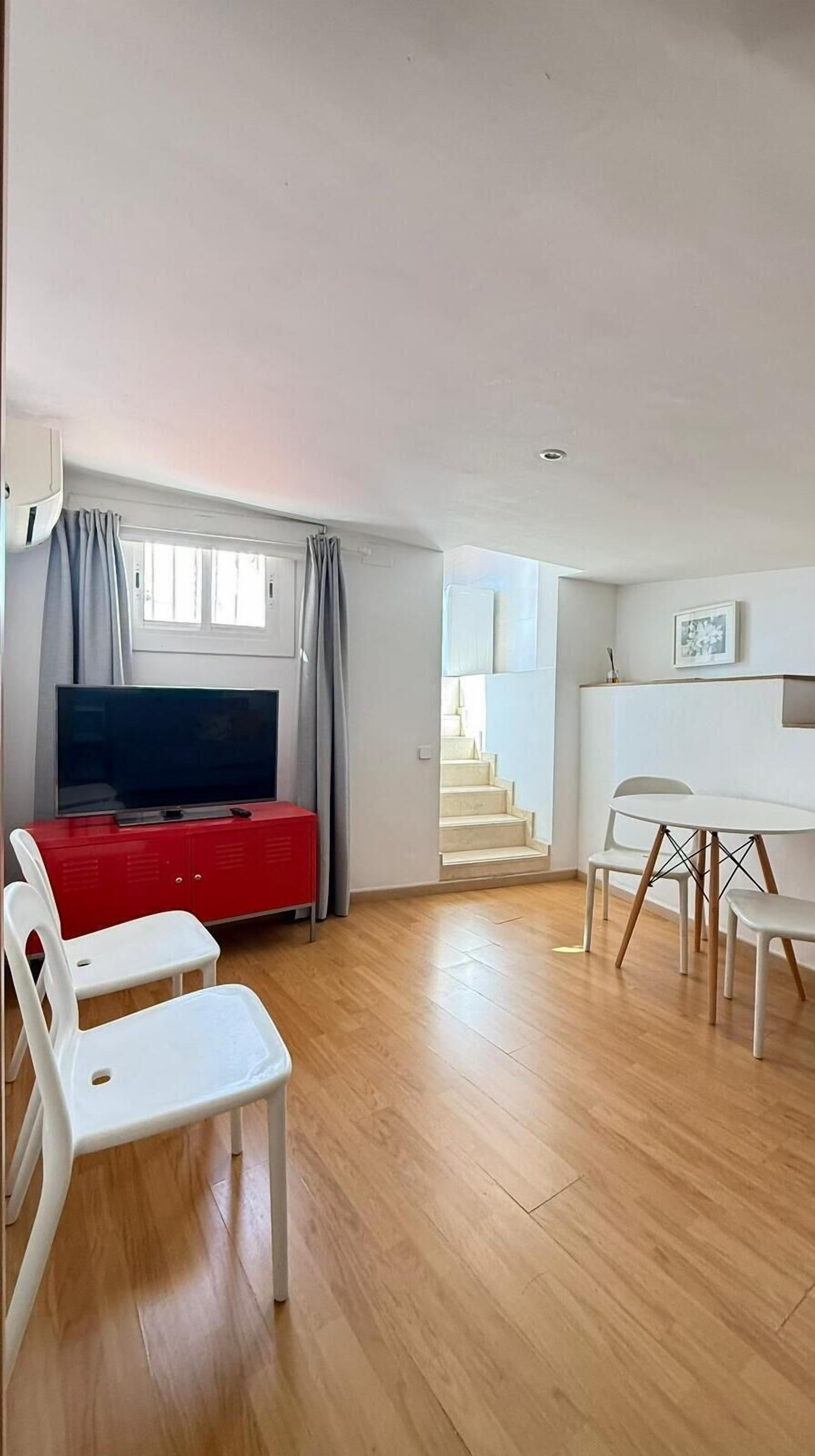 Amplia sala de estar con televisor de pantalla plana, un armario rojo y una mesa de comedor con sillas. Una escalera conduce a un nivel superior.