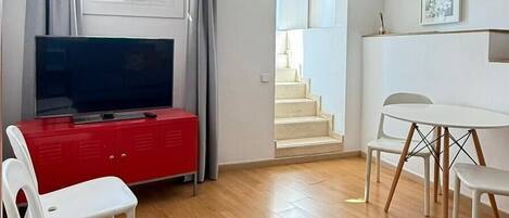 Amplia sala de estar con televisor de pantalla plana, un armario rojo y una mesa de comedor con sillas. Una escalera conduce a un nivel superior.