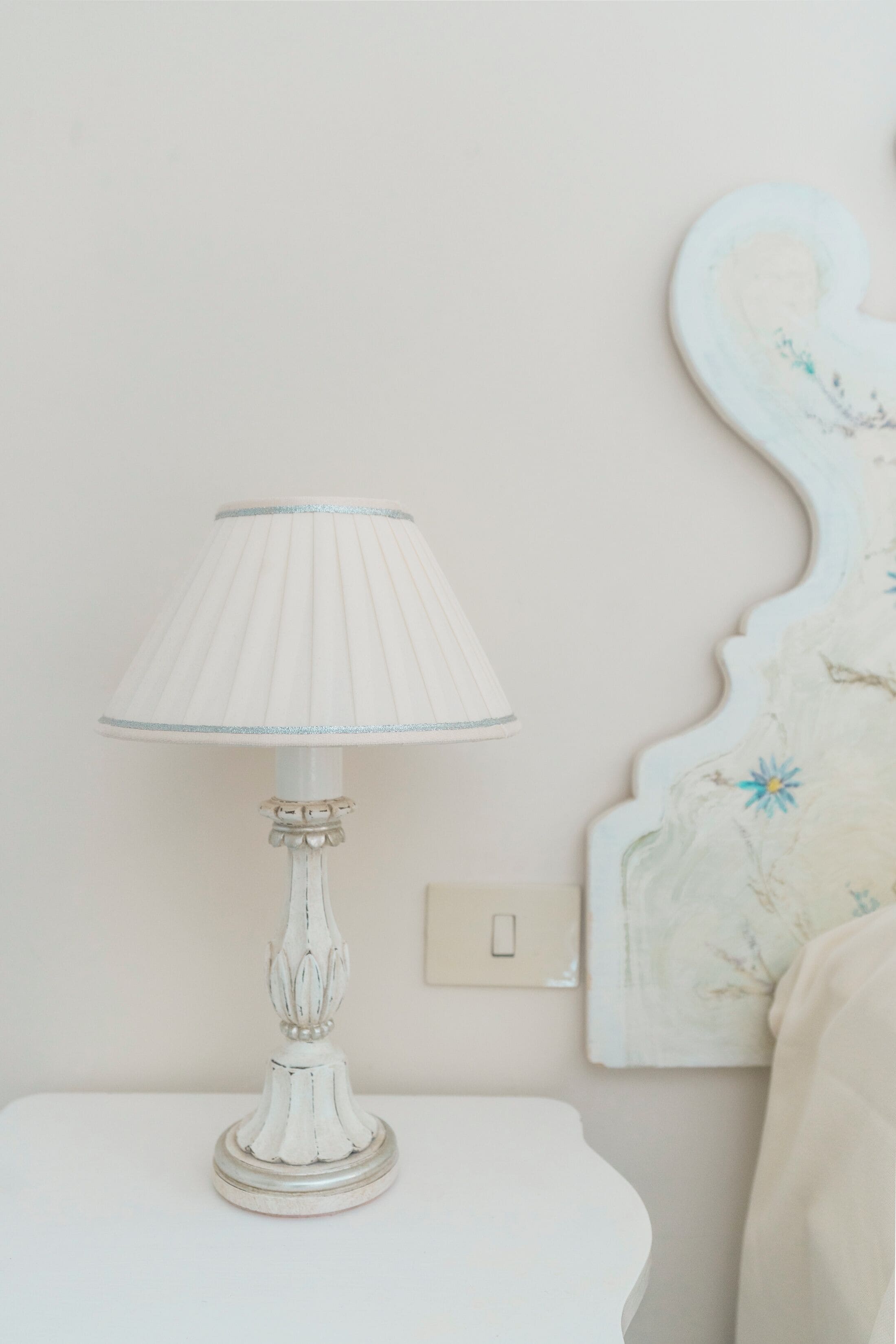 A white table lamp with a pleated shade on a white bedside table.